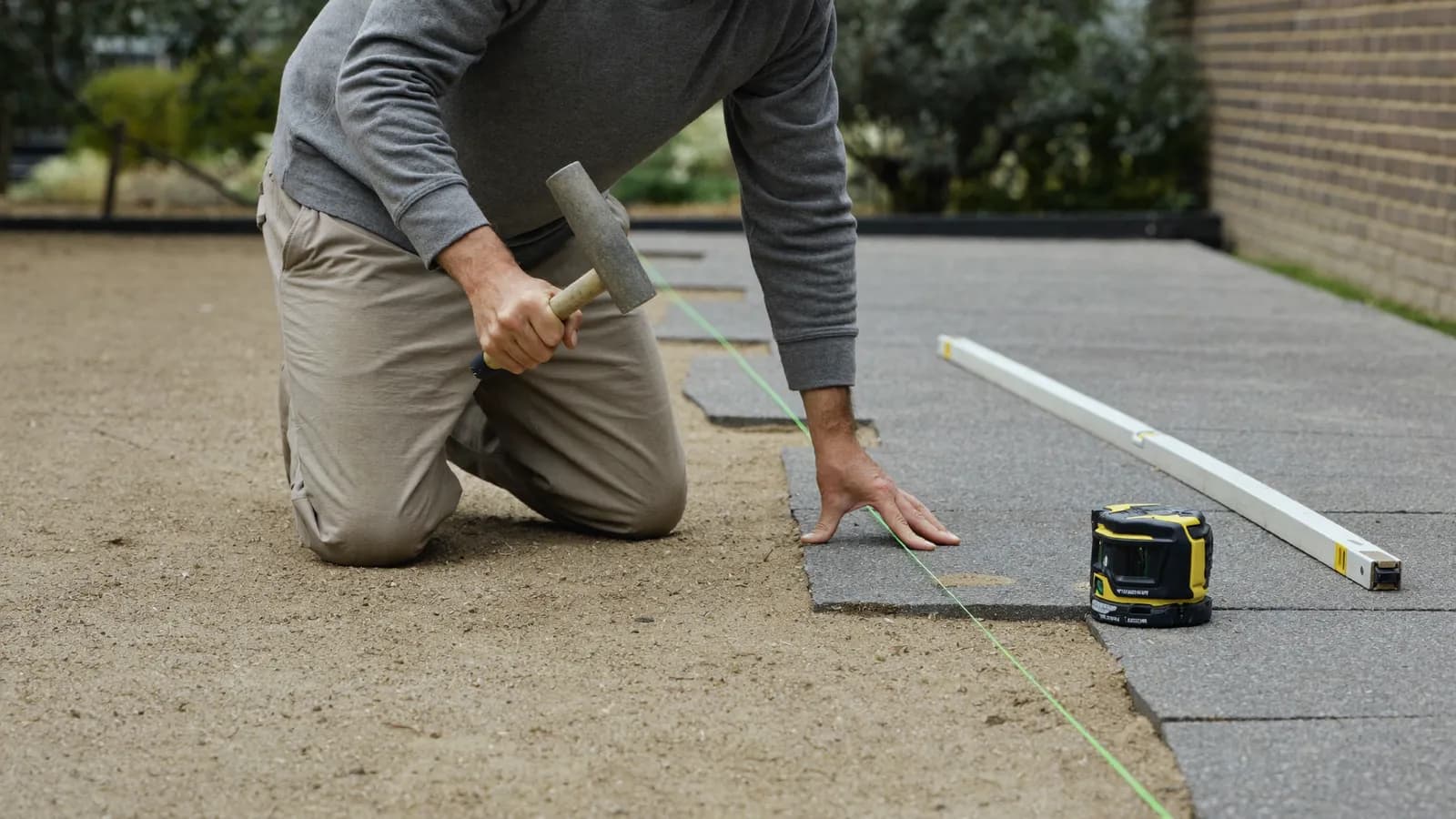 Tuinaannemer legt betonklinkers op een zandbed, met laserlijn en rijlat om het vlak uit te zetten